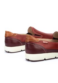Zapatos de piel marrón sin cordones con acentos en color beige, un empeine texturizado y una suela de goma blanca. Costuras visibles a lo largo de los lados, detalle sutil del logo.