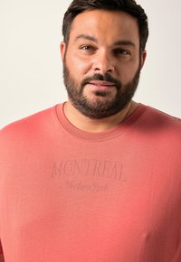 Krátký rukáv, korálová tričko s kulatým výstřihem a embosovaným textem "MONTREAL Northern Roots" na přední straně, se hladkým povrchem.