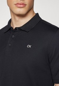 Crna polo majica od teksturirane tkanine. Ima ovratnik, tri dugmeta i mali bijeli "CK" logo na prsima.