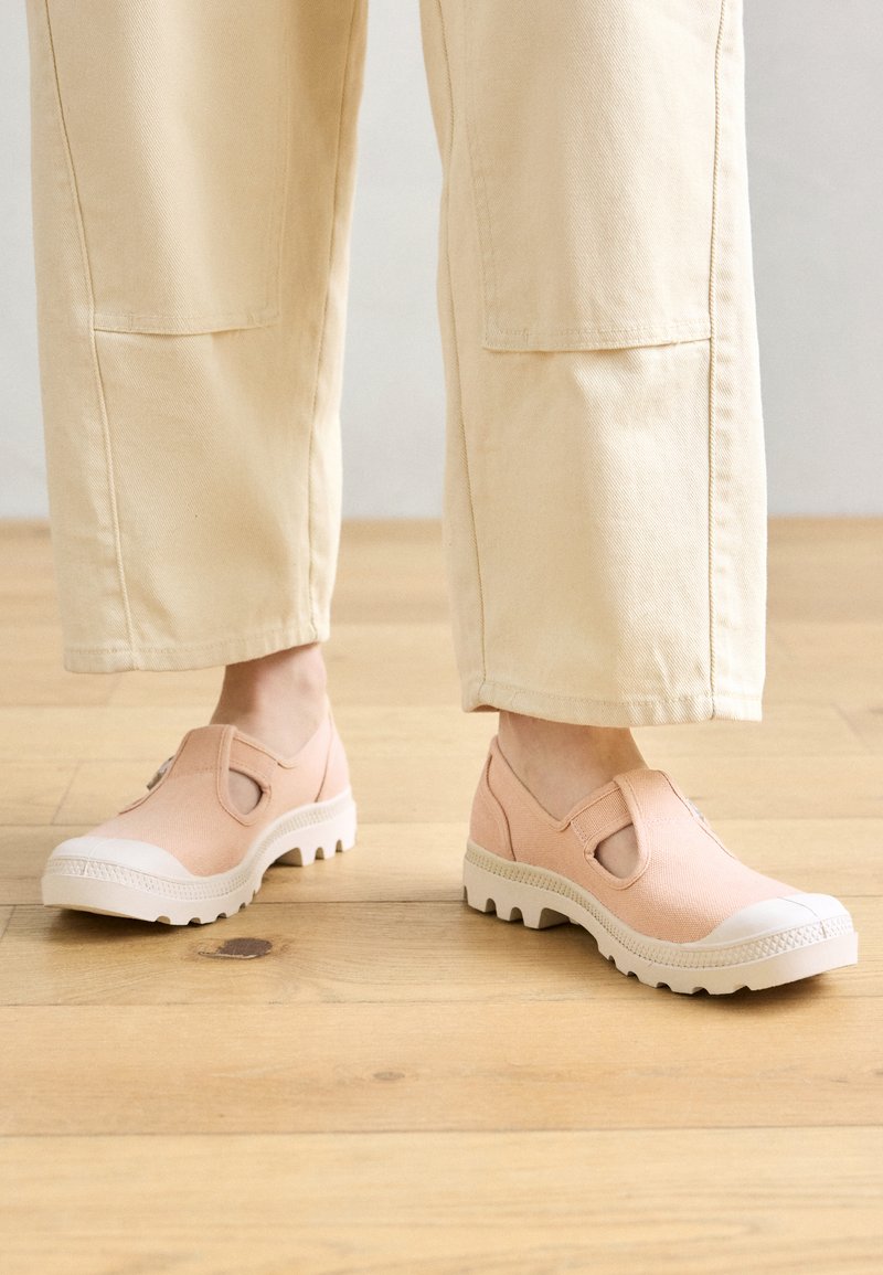 Persona che indossa pantaloni beige chiaro e scarpe slip-on in tela rosa con suole bianche, in piedi su un pavimento di legno.