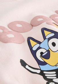 Sweat-shirt rose avec une impression en relief texturée d’un personnage de dessin animé aux accents bleus et jaunes, et de grandes lettres noires et blanches.