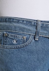 Siniset farkut, joissa on lievä kulutus, pieni etutasku CK-logolla, vyölenkit ja kontrastisauma.