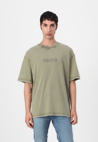 Olive green cotton t-shirt with short sleeves and a round neckline, featuring a central, grey "HUGO" logo and visible stitching details.