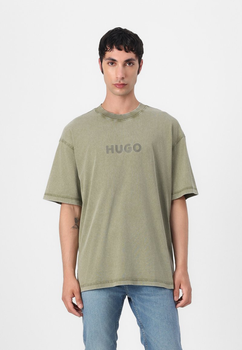 Olive green cotton t-shirt with short sleeves and a round neckline, featuring a central, grey "HUGO" logo and visible stitching details.