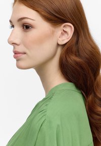 Blusa verde con una texture morbida e dettagli plissettati, indossata da una persona con lunghi capelli rossi ondulati, vista di profilo su uno sfondo bianco.