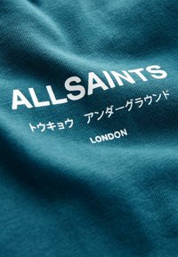 Tirkizna tkanina s teksturiranim završetkom s otiskom "ALLSAINTS" u bijeloj boji, zajedno s japanskim tekstom i "LONDON" ispod.