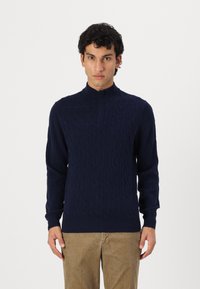 Granatowy sweter dzianinowy z wysokim kołnierzem i wzorem w warkocze, z długimi rękawami i ściągaczem u dołu. Noszony z beżowymi spodniami.