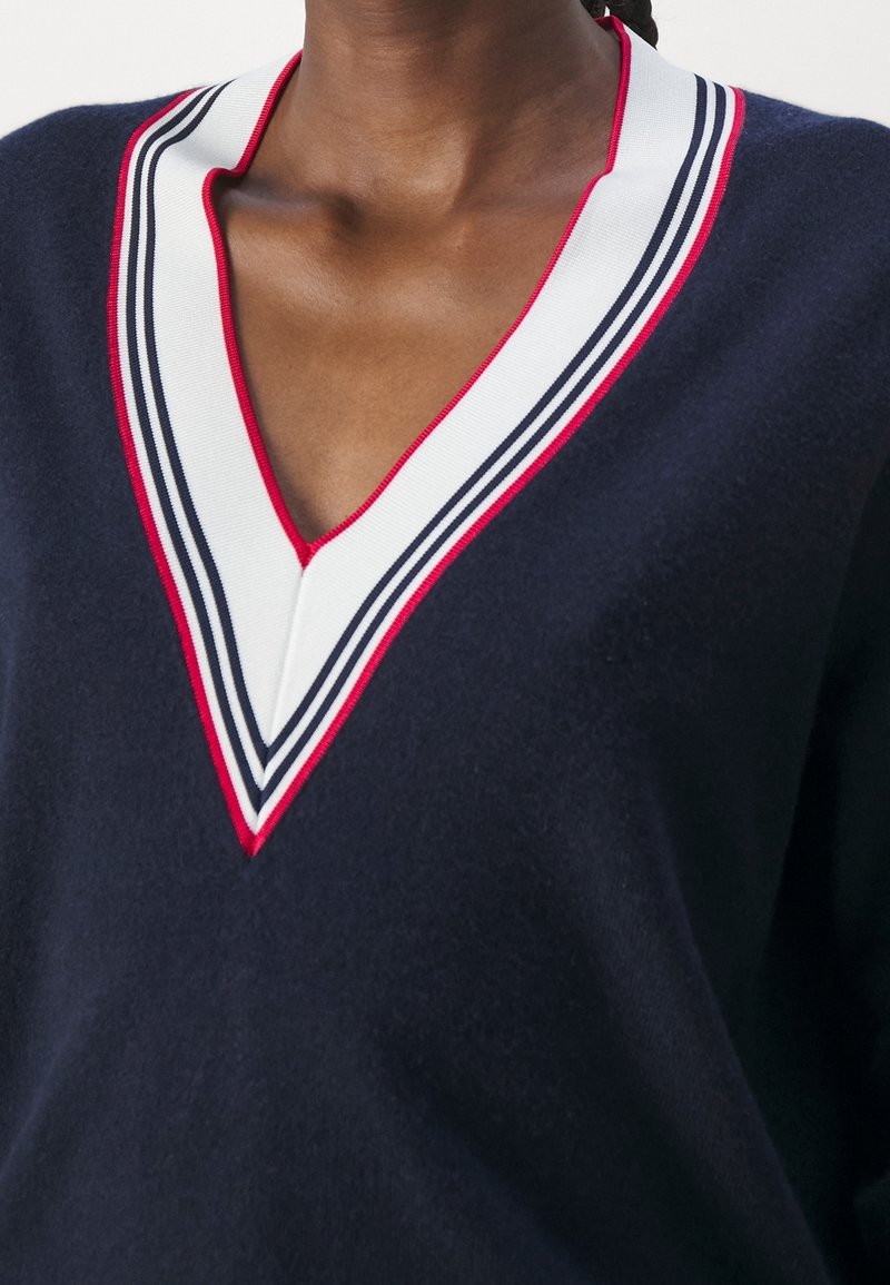 Pull bleu marine foncé à col en V avec bordure rayée blanche, bleu marine et rouge autour de l'encolure, porté par une personne à la peau foncée.