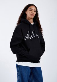 Black hooded sweatshirt made of cotton, featuring a white signature logo on the front, with a kangaroo pocket and adjustable drawstrings.