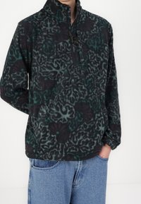 Osoba ubrana w polarową bluzę z ciemnym wzorem i zamkiem błyskawicznym na piersi, w niebieskie dżinsy, z ręką w kieszeni, stojąca na białym tle.