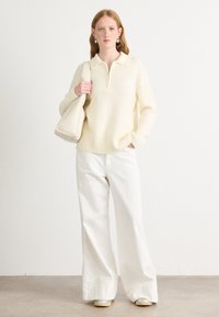 Maglione a maglia crema con colletto e chiusura a zip, abbinato a pantaloni bianchi a gamba larga e una borsa rigida crema. Scarpe argento completano l'outfit.