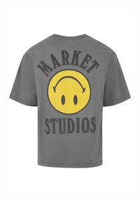 Pilkos spalvos medvilninė marškinė su dideliu geltonu šypsenėlės grafiku ir juodomis storomis raidėmis „MARKET STUDIOS“ ant nugaros.