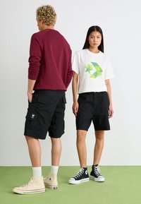 Sorte cargo-shorts med flere lommer, parret med en hvid t-shirt med grøn palme-grafik og en vinrød sweatshirt, båret med kontrasterende sneakers.