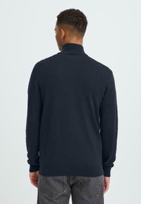 Pull en maille bleu marine avec col montant, poignets et ourlet côtelés. Texture douce et détails minimalistes au dos.