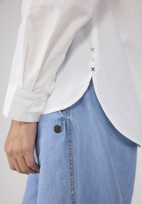 Weißes Hemd mit geknöpften Manschetten und gesteppten Akzenten in Rosa, Grau und Grün an der Seitennaht, kombiniert mit blauen Denim-Hosen mit einem Metallknopf.