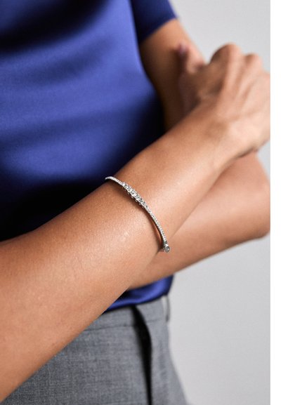 Silberarmband mit geschwungenem Design und weißen Steindetails, getragen an einem nackten Arm mit glatter Textur und dezentem Glanz.
