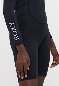 Crni neopren sa kratkim rukavima, s lavandom "ROXY" logom na ruci. Ima pripijen dizajn i glatku, rastezljivu teksturu.