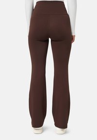 Femme portant un pantalon évasé taille haute marron avec des baskets blanches, vue de dos sur un fond blanc uni.