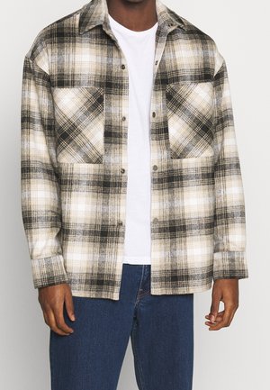 Mężczyzna ubrany w beżową, czarną i białą koszulę w kratę na guziki, na białej koszulce i niebieskich dżinsach, pokazany od klatki piersiowej do bioder.