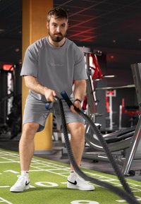 Grijze sport t-shirt en shorts met een logo, witte sneakers met zwarte accenten, bezig met battle ropes op een groene gymvloer. Fitnessomgeving.