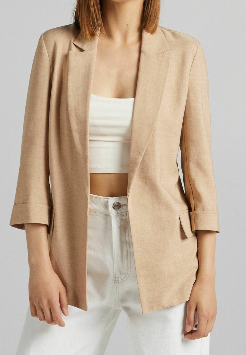 Blazer beige à coupe décontractée, avec revers crantés, poignets retroussés et deux poches avant, associé à un haut blanc et un jean clair.