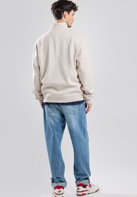 Pullover beige con colletto alto, tessuto morbido e vestibilità rilassata. Indossato con jeans azzurri e sneaker bianche-rosse, mostrato di schiena.