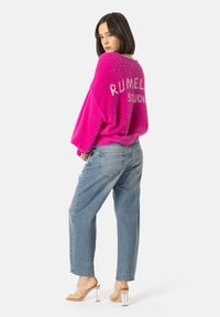 Flauschiger rosa Pullover mit "RUMEL" und "STUDIO" in Weiß auf dem Rückenteil, kombiniert mit hellblauen, lässigen Jeans und klaren Absatzsandalen.