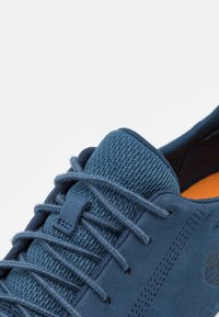 Zapato azul de gamuza con parte superior de malla texturizada, cordones redondos y un forro interior naranja. Presenta un logo embossado en el costado.