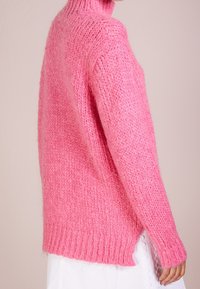 Różowy sweter z dzianiny z wysokim kołnierzem, ściągaczami przy rękawach i bocznymi rozcięciami. Miękka tekstura i luźny krój, eksponujący grubą przędzę.