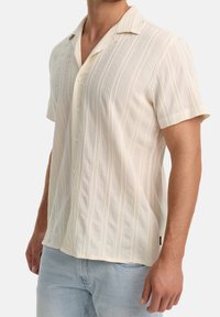 Homme portant une chemise crème à manches courtes texturée avec boutons et un jean bleu clair, debout devant un fond uni.