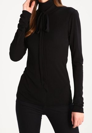 Femme portant un haut noir à manches longues et col roulé avec un détail de nœud noué au niveau du cou et un pantalon noir ajusté.