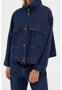 Person som bär en mörkblå denimjacka med stora framfickor och tryckknappar, kombinerad med matchande denimbyxor.