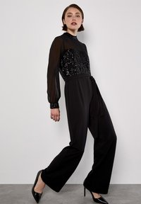 Schwarzer Jumpsuit mit einem mit Pailletten verzierten Oberteil, transparenten langen Ärmeln, einer taillierten Taille mit Gürtel und weitem Bein.