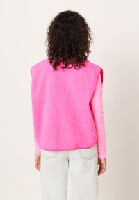 Flis telovnik v svetlo roza barvi z teksturirano površino, brez rokavov in z zaobljenim robom, kombiniran z dolgimi roza majico in svetlimi kavbojkami.