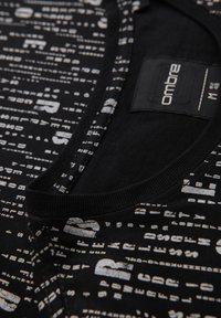 Camiseta de algodón negra que presenta un patrón texturizado de letras y números blancos. Cuello redondo y etiqueta con el nombre de la marca visible.