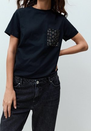 Frau trägt ein schwarzes T-Shirt mit kurzen Ärmeln und einer gemusterten Tasche auf der linken Brust sowie hoch taillierte schwarze Jeans.