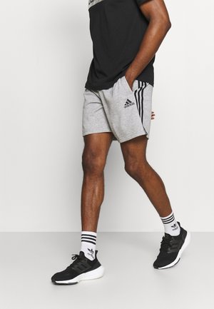 Man loopt en draagt grijze Adidas short, zwarte Adidas sneakers, witte Adidas sokken en een zwart T-shirt tegen een effen witte achtergrond.