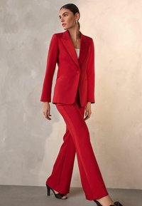 Rød skræddersyet blazer med skarpe klapper og en enkelt knap, parret med bukser med vidde, der har en kontrasterende sidelinje. Glat stoftekstur.