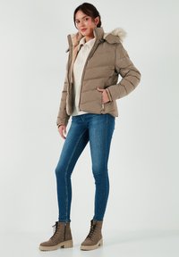 Giacca trapuntata beige con cappuccio foderato in pelliccia, fronte con zip e tasche laterali. Abbinata a un maglione crema e jeans skinny blu. Stivali marroni con lacci.