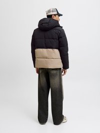 Juodos ir smėlinės spalvos pamušalu striukė su kapišonu; turi stilingą dizainą ir tekstūruotą audinį. Pora su plačiomis tamsiomis džinsomis ir sportbačiais.