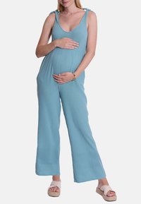 Hellblauer, ärmelloser Jumpsuit mit Bindebändern, gerippter Textur und weitem Bein-Design. Das Modell hält einen schwangeren Bauch und trägt Sandalen.