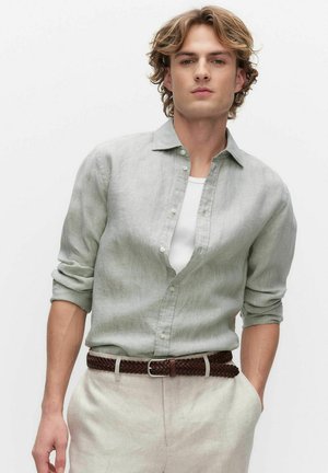 Nuori mies kiharatukka, joka käyttää vaaleanharmaata napillista paitaa valkoisen t-paidan päällä, panta sisään vaaleanbeigeihin housuihin ja ruskea punottu vyö.