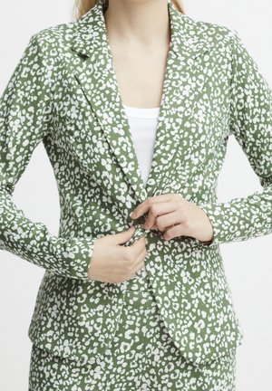 Femme boutonnant un blazer vert à taches blanches abstraites, portant un pantalon assorti et un haut blanc en dessous.