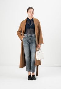 Donna che indossa un cappotto cammello sopra una maglia trasparente blu navy con un reggiseno, jeans grigi, mocassini neri e tiene in mano una piccola borsa bianca.