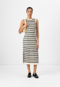 Robe en tricot avec des rayures horizontales noires et crème, un col rond, une fente sur le côté, associée à des mocassins noirs et un sac à main beige.