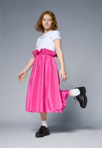 White t-shirt with logo, paired with a pink satin skirt featuring a ruffled waistband. Black chunky shoes and white crew socks.