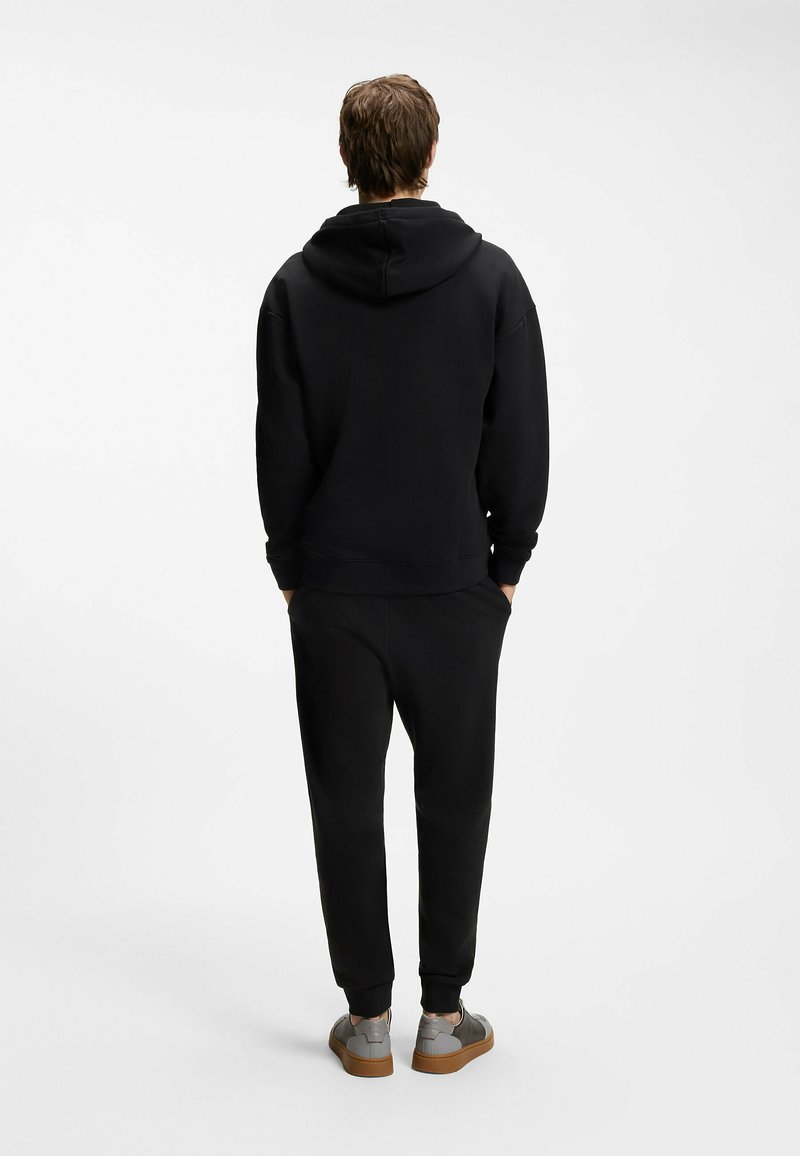Personne debout, dos tourné au spectateur, portant un sweat à capuche noir, un pantalon noir et des baskets grises avec des semelles marron.