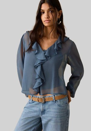 Blue sheer blouse with ruffled V-neckline, long sleeves, and a loose fit. Paired with high-waisted light denim jeans and a tan belt.
