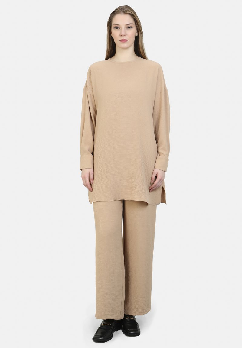 Set composto da un tunica beige a maniche lunghe e pantaloni larghi. Tessuto morbido e testurizzato con fessure laterali; abbinato a scarpe nere.