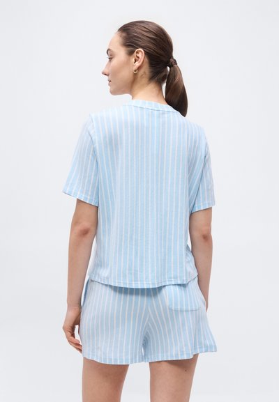 Γυναίκα με καστανά μαλλιά σε αλογοουρά, φοράει κοντομάνικη μπλούζα με κάθετες ρίγες σε ανοιχτό μπλε και λευκό και ταιριαστά σορτς, στέκεται με την πλάτη προς τα εμπρός.